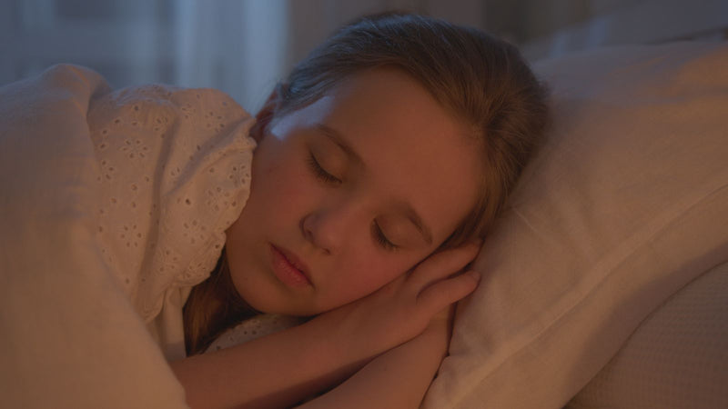 A young child asleep in bed in a softly lit room, covers pulled up and face relaxed