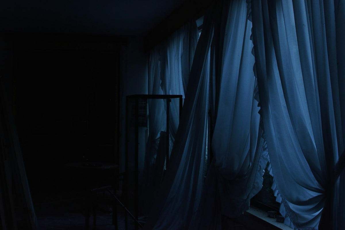 A dimly lit bedroom with sheer curtains drawn across the window and a chair beside the bed